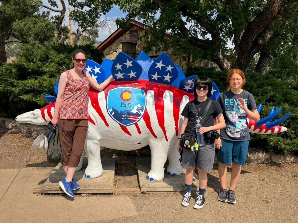 A family of three stands beside a large stegosaurus statue painted with red and white stripes and blue stars resembling the American flag. The dinosaur features a Colorado state seal on its side.