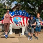 A family of three stands beside a large stegosaurus statue painted with red and white stripes and blue stars resembling the American flag. The dinosaur features a Colorado state seal on its side.