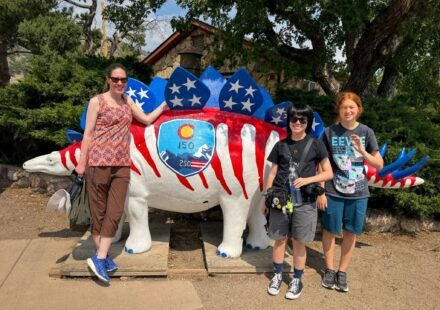 A family of three stands beside a large stegosaurus statue painted with red and white stripes and blue stars resembling the American flag. The dinosaur features a Colorado state seal on its side.