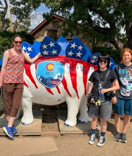 A family of three stands beside a large stegosaurus statue painted with red and white stripes and blue stars resembling the American flag. The dinosaur features a Colorado state seal on its side.