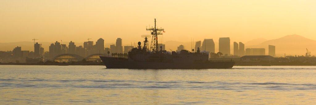A ferry sailing on calm waters with a city skyline and mountain in the background during sunset. The warm colors create a peaceful and scenic view.