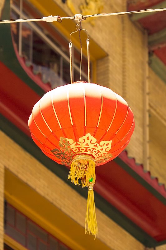 A vibrant red lantern hangs gracefully from a string, adorned with golden details and a yellow tassel, against an ornate architectural backdrop.