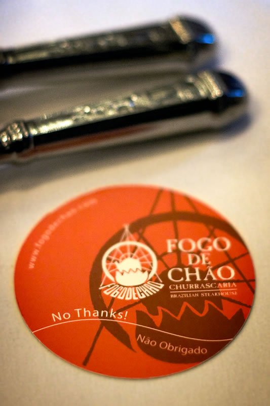 A close-up image shows an orange Ffogo de Chão coaster with 'No Thanks!' written on it, next to two elegant black pens.