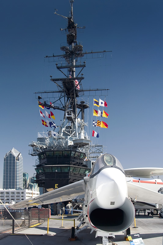 A fighter jet flies low over an aircraft carrier deck with a clear blue sky in the background.