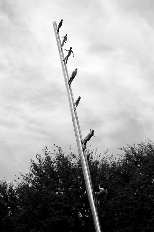 A black and white photo of multiple figures climbing a pole against a cloudy sky.
