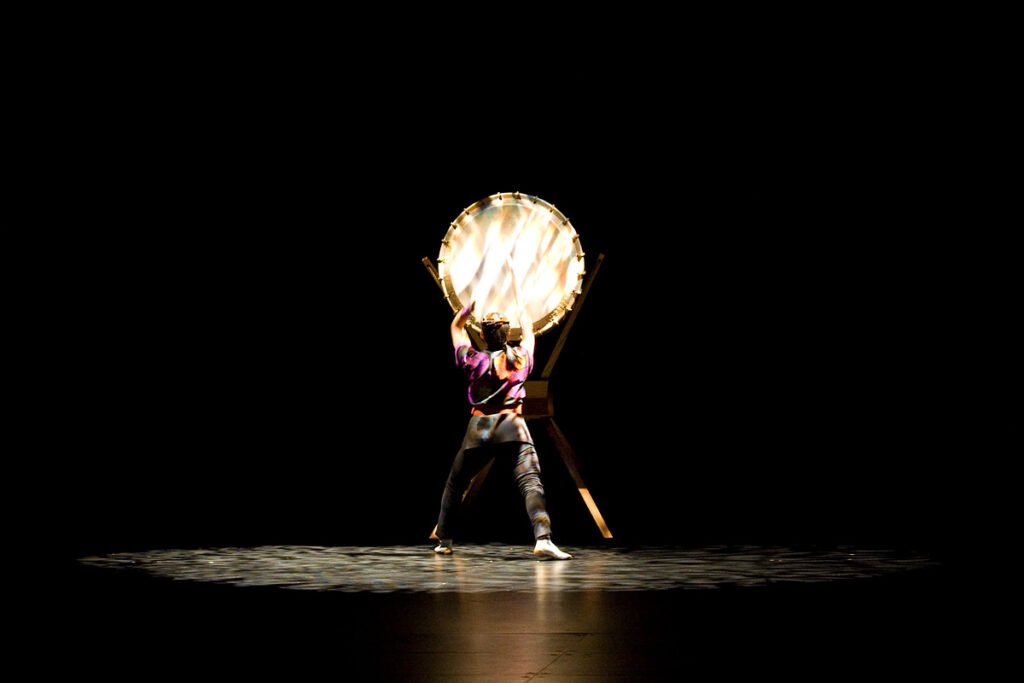 A dancer on stage wearing a fire-breathing mask, illuminated against the dark background.