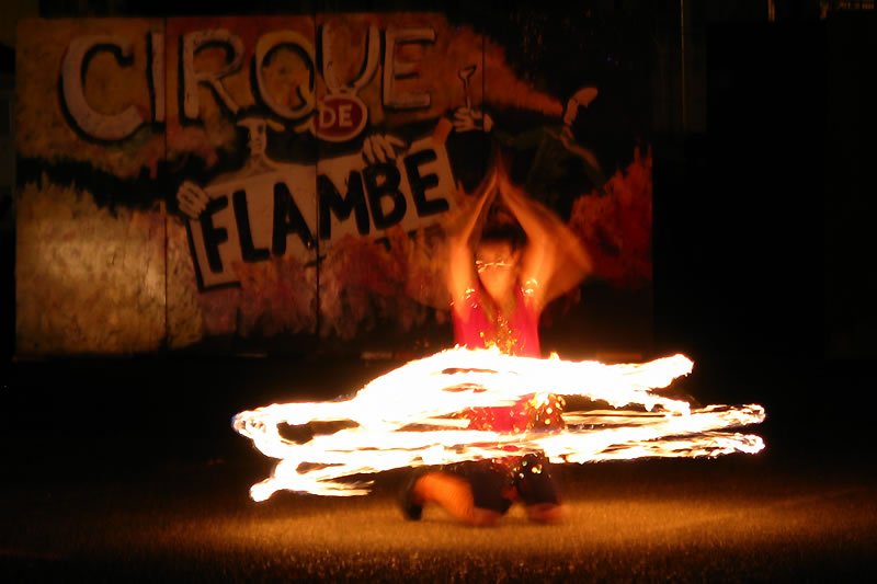 A fire dancer performs in front of a sign reading 'Cirque de Flambe' at night. Bright flames swirl around her as she dances barefoot on grass.