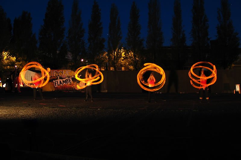 Four performers spin glowing orange hula hoops under a deep blue night sky.
