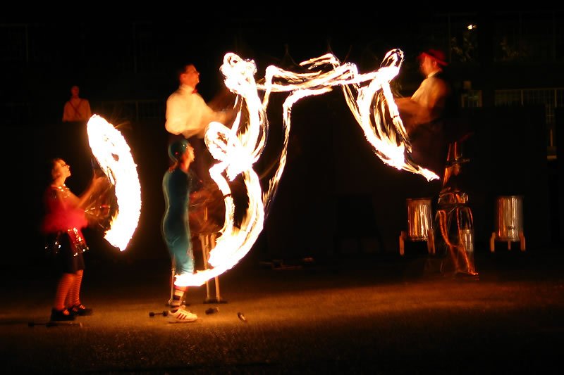 A group of performers engage in a fire dance under the night sky, creating intricate patterns with flames. The scene is illuminated by the bright, swirling lights against a dark background.