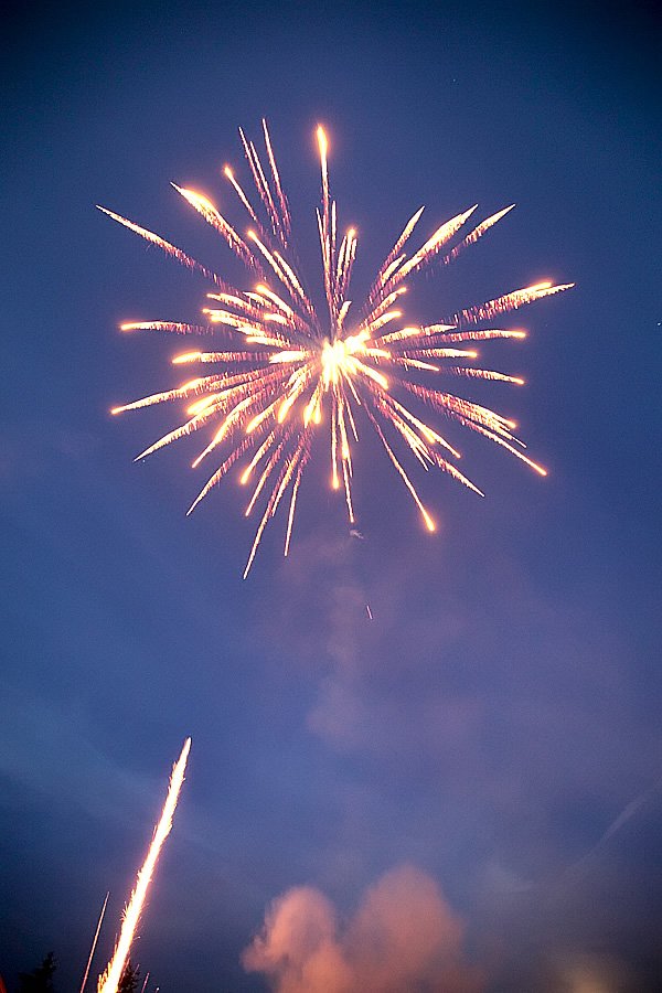Vibrant fireworks explode in golden bursts against a deep blue sky, with soft clouds glowing below.