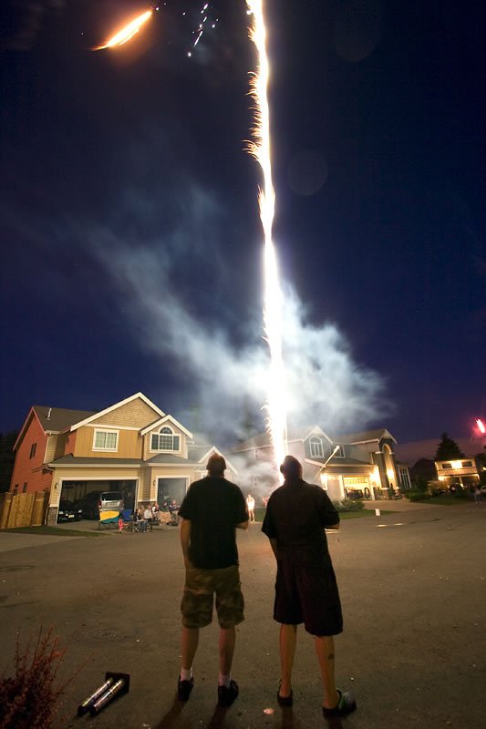 A vibrant display of fireworks exploding in the twilight sky, with streaks of golden light radiating outward. A single firework ascends toward the center, leaving a bright trail against the deep blue dusk.
