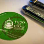 Dallas A green Fogo de Chão coaster with the text 'Yes Please!' and two shiny saltshakers on a white surface.