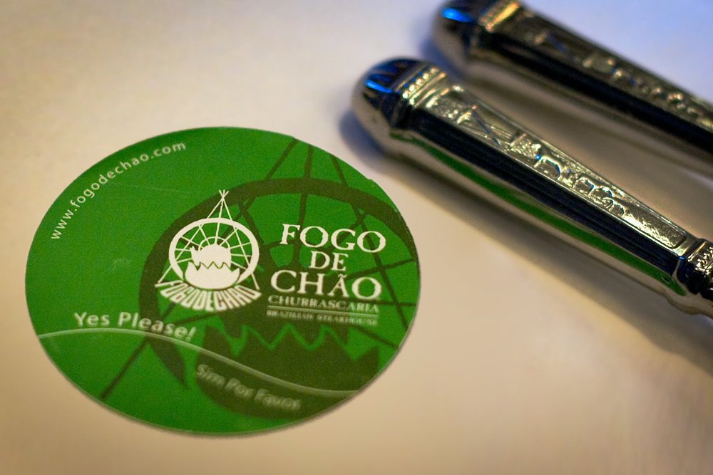 A green Fogo de Chão coaster with the text 'Yes Please!' and two shiny saltshakers on a white surface.