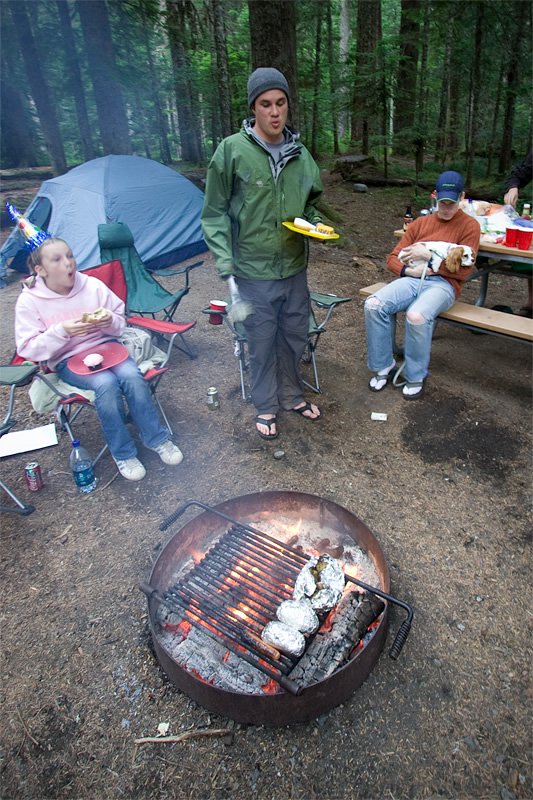 People gather around a fire pit in a forest setting, preparing food while others watch.