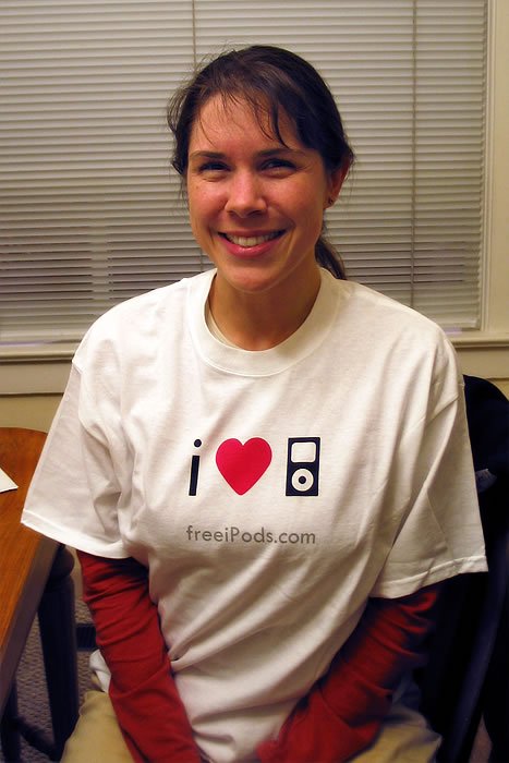 Kristie smiling while wearing a white t-shirt with an iPod graphic. A red long-sleeve is visible underneath and blinds are in the background.