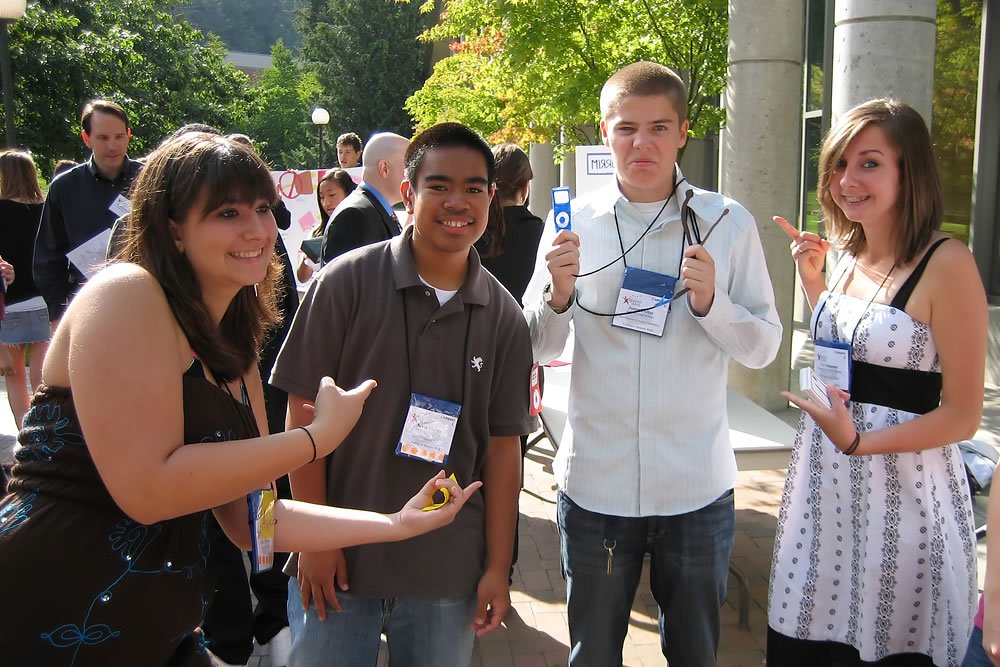 A group of four friends smiling and posing together at an outdoor conference event. They are wearing lanyards and enjoying each other's company in a casual setting.
