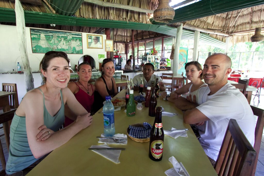 A group of friends sitting around a long wooden table under a thatched roof, enjoying food and drinks together.