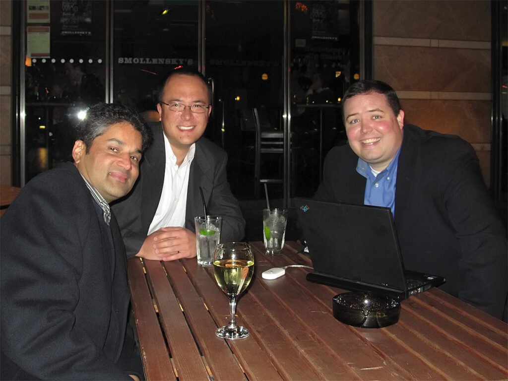 Three men enjoying drinks and conversation at a wooden table, with one using a laptop.