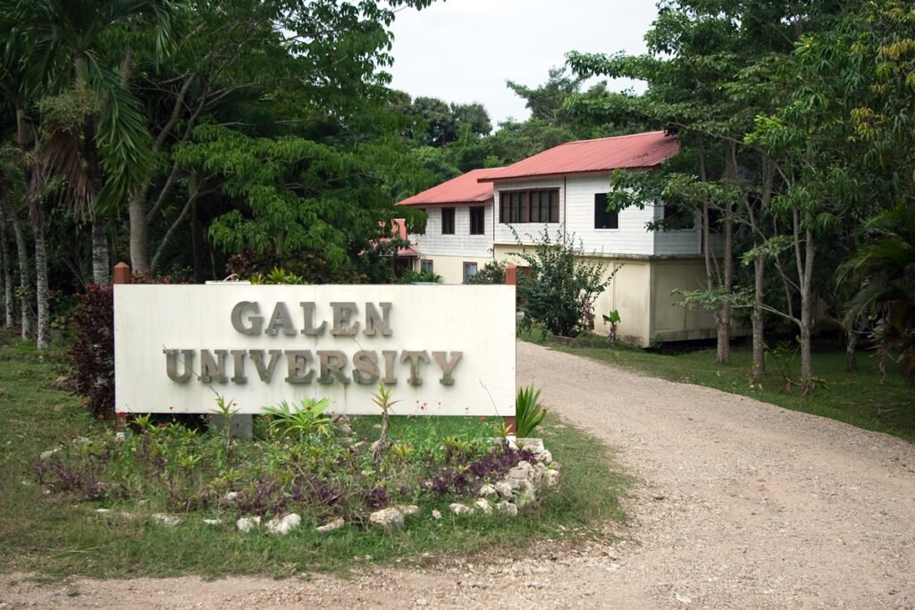 A dirt road leads to a white building with a red roof, marked by a sign reading 'Galen University' surrounded by lush greenery.