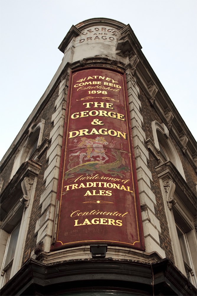 Ornate Victorian-era pub facade with a burgundy banner reading 'The George & Dragon' advertising Watney's ales. Weathered stone architecture and classic English pub styling characterize this historic establishment.