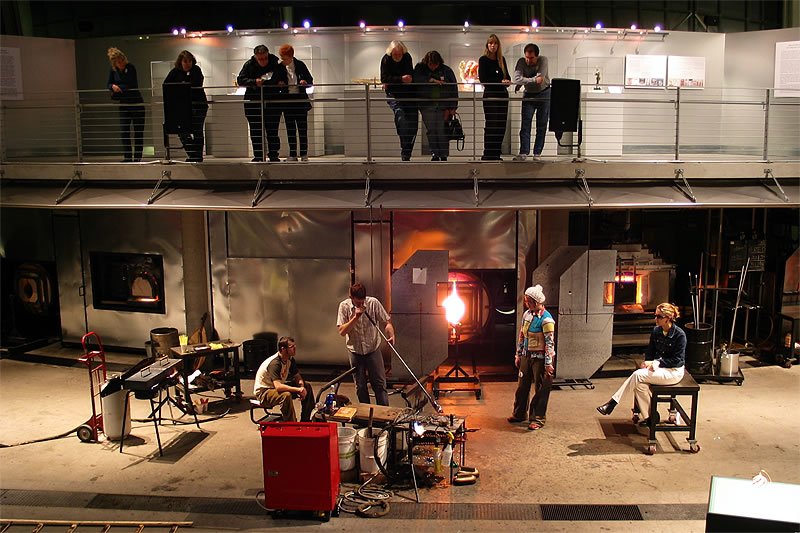 Artists work at a hot glass station below an upper-level viewing gallery.