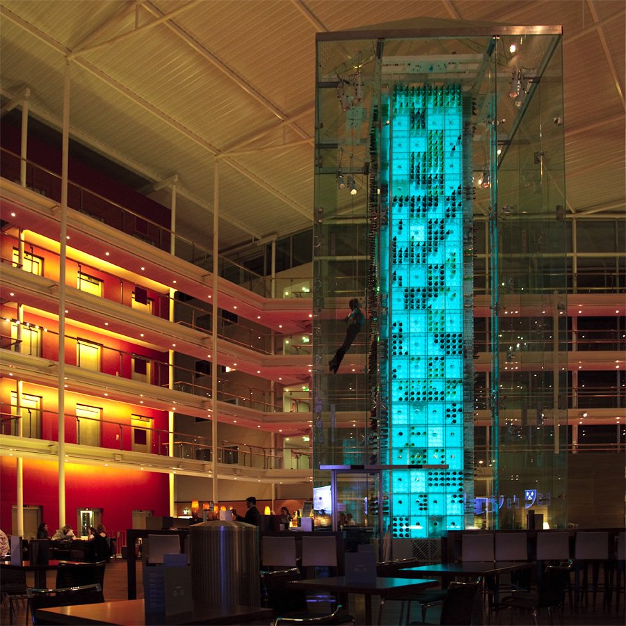 A striking glass elevator with blue lighting stands tall in a multi-level hotel atrium featuring warm red and yellow tones.