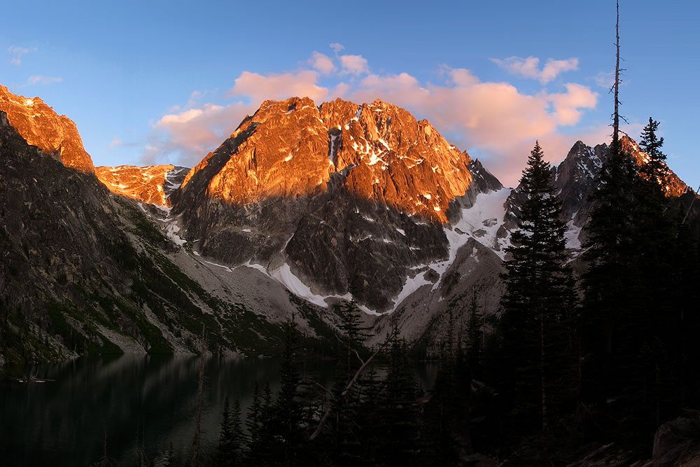 Sunlit mountain peaks with snow patches. Silhouetted evergreen trees in foreground.
