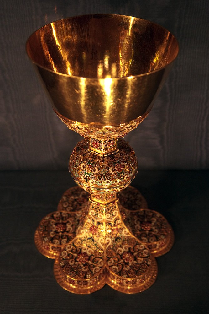 A golden chalice with intricate floral patterns on its base stands prominently against a dark background.