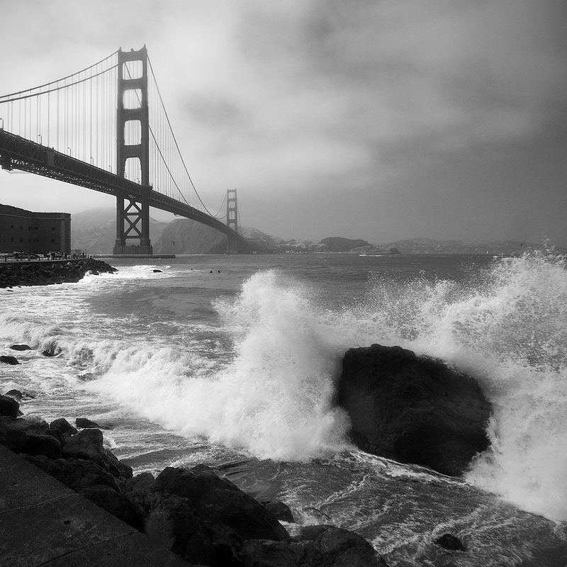 A dramatic black-and-white image of the Golden Gate Bridge with powerful waves crashing against rocks.