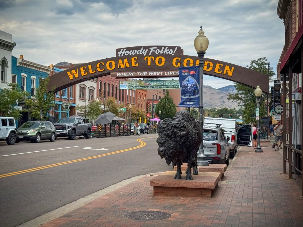 A picturesque street in Golden, Colorado, featuring a welcoming sign and a prominent bison statue. The scene is set against a backdrop of historic buildings and mountains.