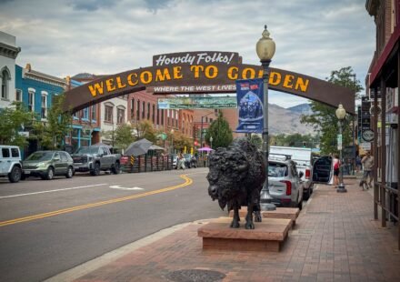 A picturesque street in Golden, Colorado, featuring a welcoming sign and a prominent bison statue. The scene is set against a backdrop of historic buildings and mountains.