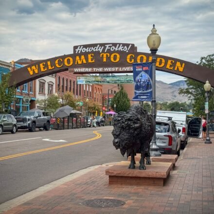 A picturesque street in Golden, Colorado, featuring a welcoming sign and a prominent bison statue. The scene is set against a backdrop of historic buildings and mountains.