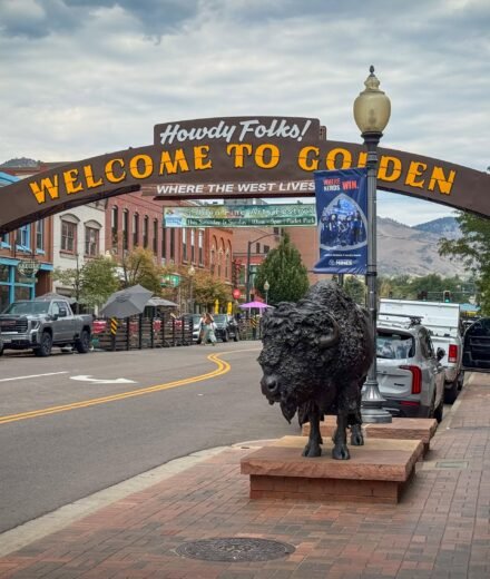 A picturesque street in Golden, Colorado, featuring a welcoming sign and a prominent bison statue. The scene is set against a backdrop of historic buildings and mountains.
