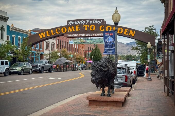 A picturesque street in Golden, Colorado, featuring a welcoming sign and a prominent bison statue. The scene is set against a backdrop of historic buildings and mountains.