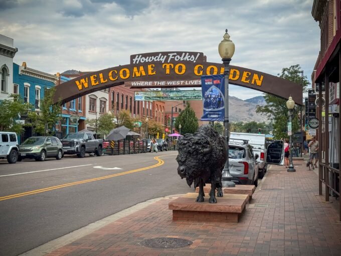 A picturesque street in Golden, Colorado, featuring a welcoming sign and a prominent bison statue. The scene is set against a backdrop of historic buildings and mountains.