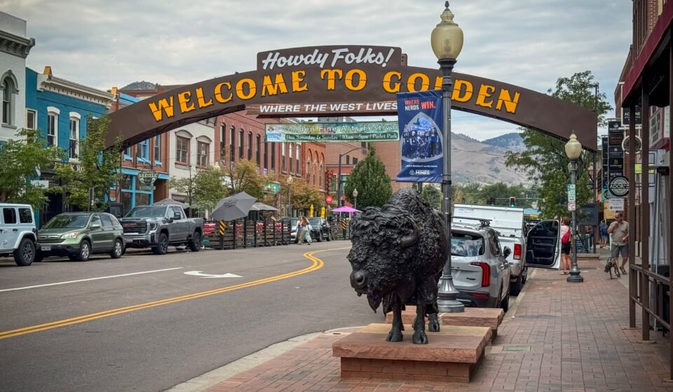 A picturesque street in Golden, Colorado, featuring a welcoming sign and a prominent bison statue. The scene is set against a backdrop of historic buildings and mountains.