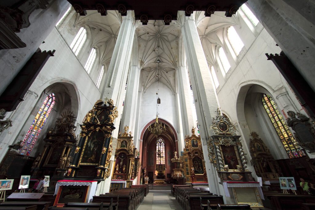 A grand church interior featuring tall Gothic arches, intricate ceiling vaults, and ornate altars adorned with religious statues and stained glass.
