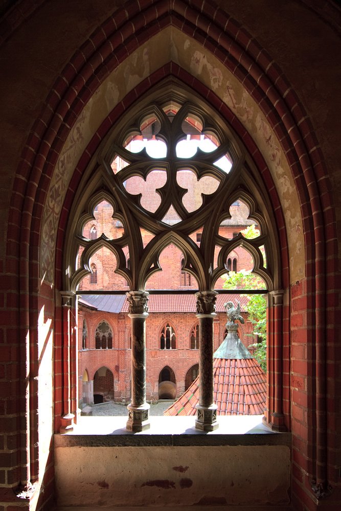 A gothic window with intricate tracery and stained glass panels, opening to a view of a courtyard with red brick buildings and greenery.