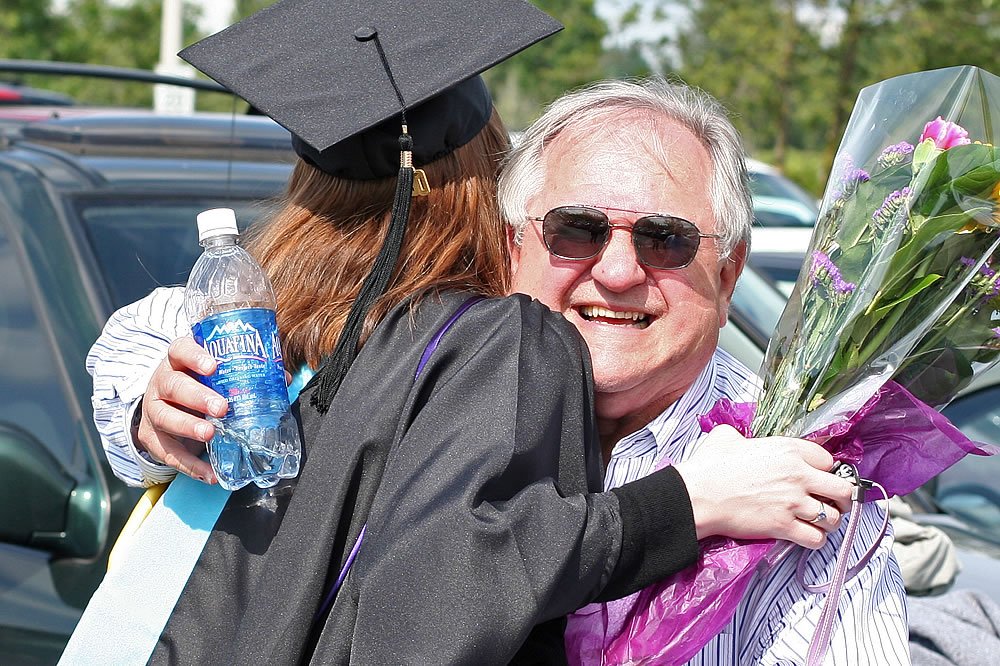 A graduate in cap and gown shares a joyful hug with an older man holding flowers. The scene captures a celebratory moment outside, possibly after graduation.