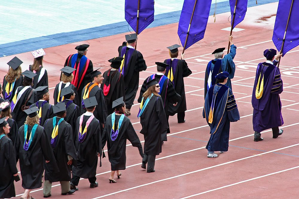 Graduates and faculty march on a running track during commencement ceremony. Colorful stoles and purple flags add vibrancy to the scene.