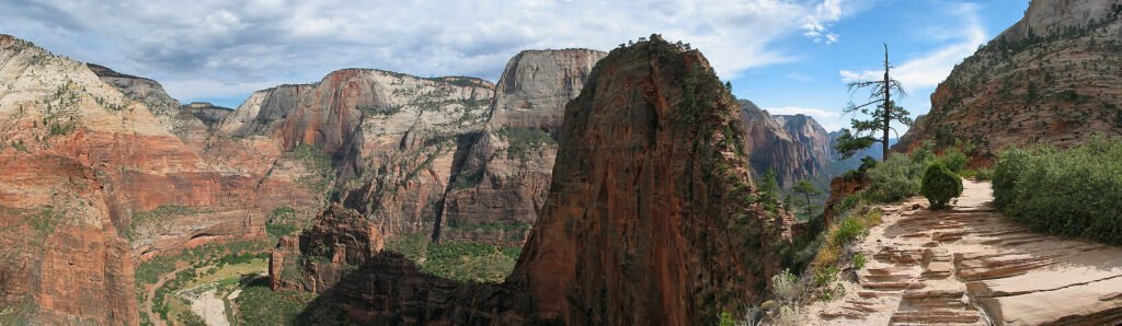 A breathtaking view of a hiking trail winding through the rugged cliffs of the Grand Canyon. The path is set among towering rock formations and lush greenery, leading towards distant mountains under a partly cloudy sky.