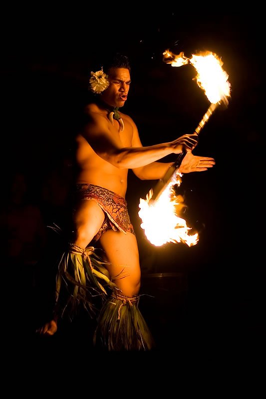 A shirtless male dancer performs with a flaming stick. He is wearing traditional grass skirt and headpiece.