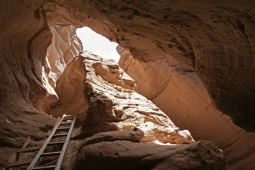 A wooden ladder leads into a narrow cave opening carved into reddish-brown rock walls.