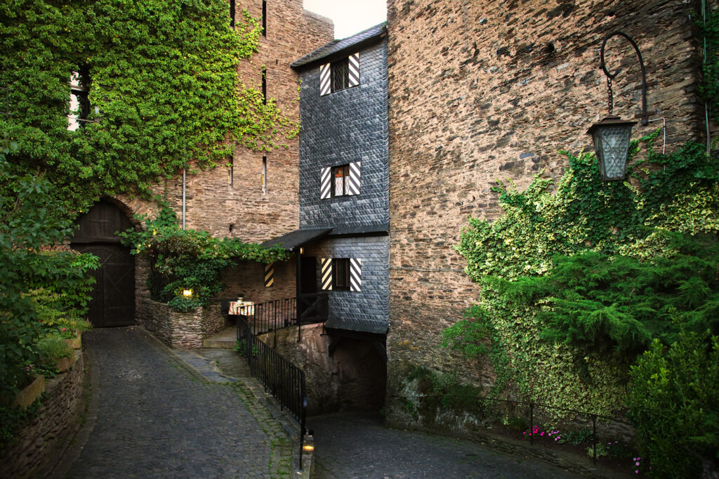 A narrow alleyway flanked by stone walls, adorned with greenery and an old-style lantern.