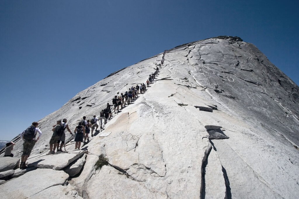 A group of hikers climbs up a steep, exposed granite slant under a clear blue sky.