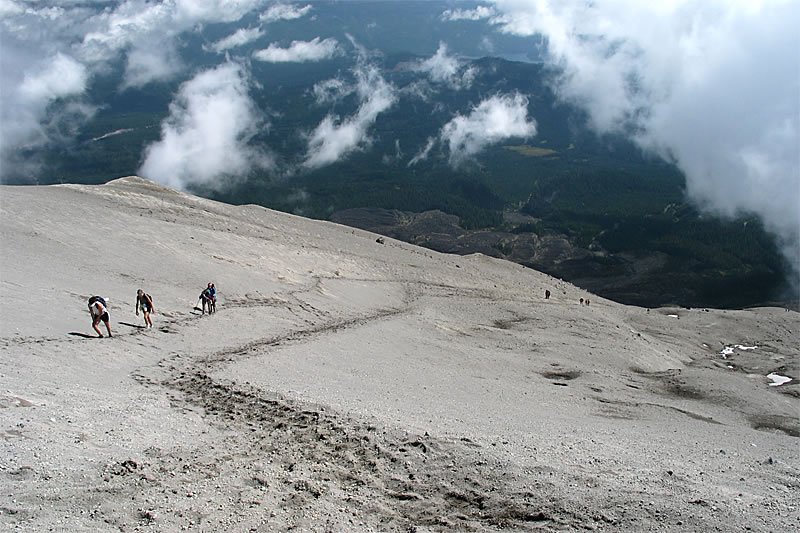 Three hikers trek along a rugged mountain trail, dwarfed by the vast landscape of clouds and distant forests below.