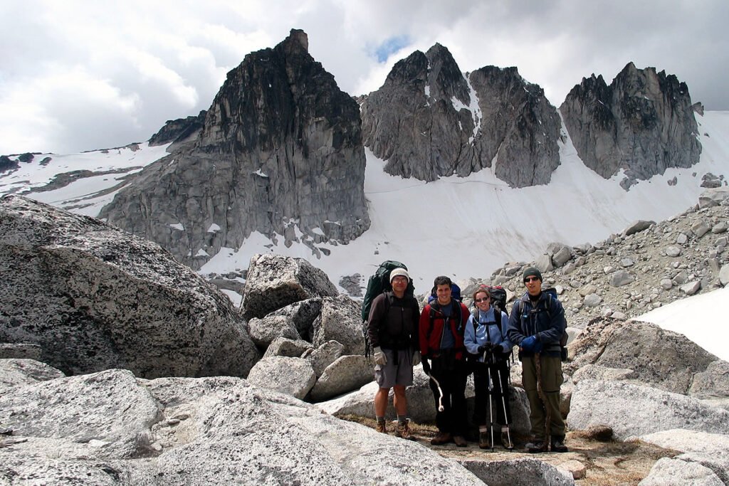 A group of four hikers standing on a rocky mountain peak with snow and dramatic peaks in the background.