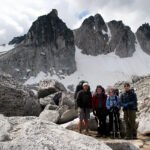 A group of four hikers standing on a rocky mountain peak with snow and dramatic peaks in the background.
