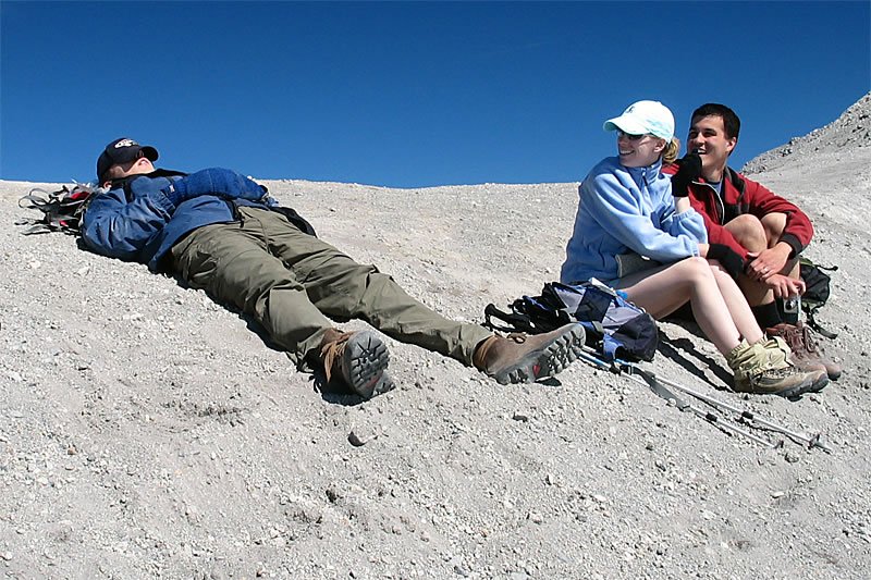 Three hikers relaxing on a rocky mountain slope. One lies down while two sit together, enjoying the sunny Pacific Northwest landscape.