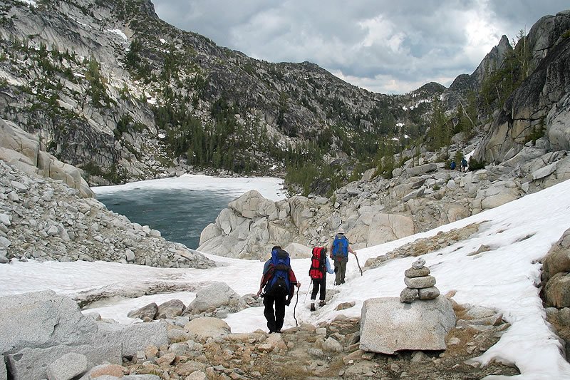 Three hikers walk along a rocky, snow-lined mountain path near a serene alpine lake, with rugged peaks rising in the background.